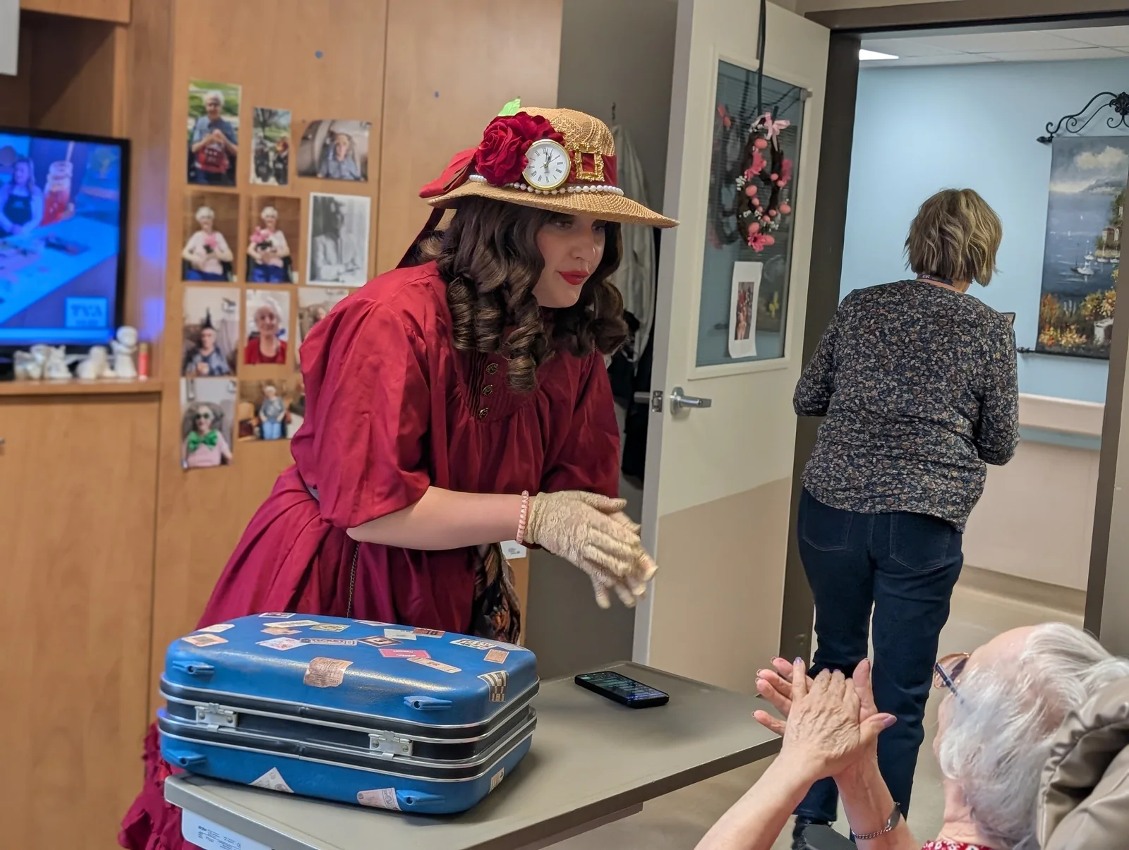 Mlle Rose-Souvenir en costume bordeaux, avec sa valise, à la rencontre d’une résidente.